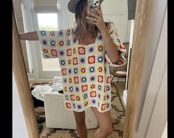 Könnte beinhalten: Eine Frau trägt ein weißes Häkelkleid mit einem bunten geometrischen Muster. Das Kleid hat einen quadratischen Ausschnitt und kurze Ärmel. Die Frau steht vor einem Spiegel und trägt einen Strohhut.