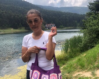 May include: A woman is holding a purple and pink crocheted tote bag with a square pattern. The bag has a light pink and white trim. The woman is standing in front of a lake with a house in the background.