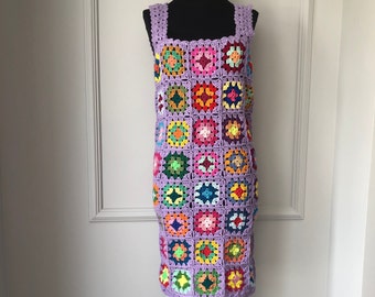 Puede incluir: Un vestido de crochet sin mangas con escote cuadrado. El vestido está hecho de hilo de color lila claro y presenta un patrón geométrico colorido de cuadrados.