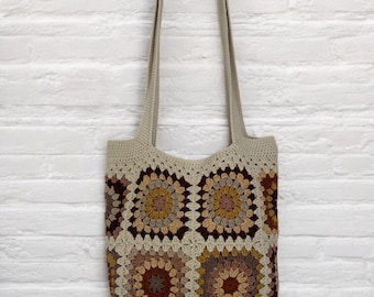 May include: A crocheted tote bag with a brown, beige, and orange geometric pattern. The bag has two long straps.