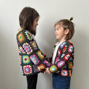 May include: Two children wearing colourful crocheted cardigans with a granny square pattern. The cardigans are black with multi-coloured squares. The children are holding hands and smiling at each other.