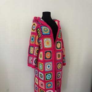 Puede incluir: Una larga chaqueta de punto de ganchillo rosa con capucha. La chaqueta está hecha de cuadrados coloridos con una variedad de patrones.