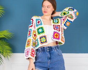 May include: A white crochet cardigan with colorful squares. The cardigan has a V-neck and is cropped. The model is wearing blue denim shorts.