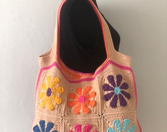 May include: A crocheted tote bag with a floral design. The bag is made of beige yarn and features six colorful flowers in shades of orange, yellow, pink, purple, blue, and green.