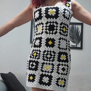 May include: A handmade crochet dress in a square pattern of black and white with yellow accents. The dress has a square neckline and is knee-length. The dress is made of yarn and is a summer garment.