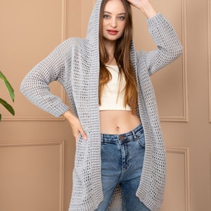 May include: A grey open-front cardigan with a hood and a loose, open knit. The cardigan is worn over a white tank top and blue jeans.