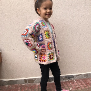 May include: A young person wearing a colorful crocheted cardigan with a fuzzy texture. The cardigan features a variety of colors, including pink, blue, green, yellow, and orange. The person is wearing black leggings and pink and green shoes.