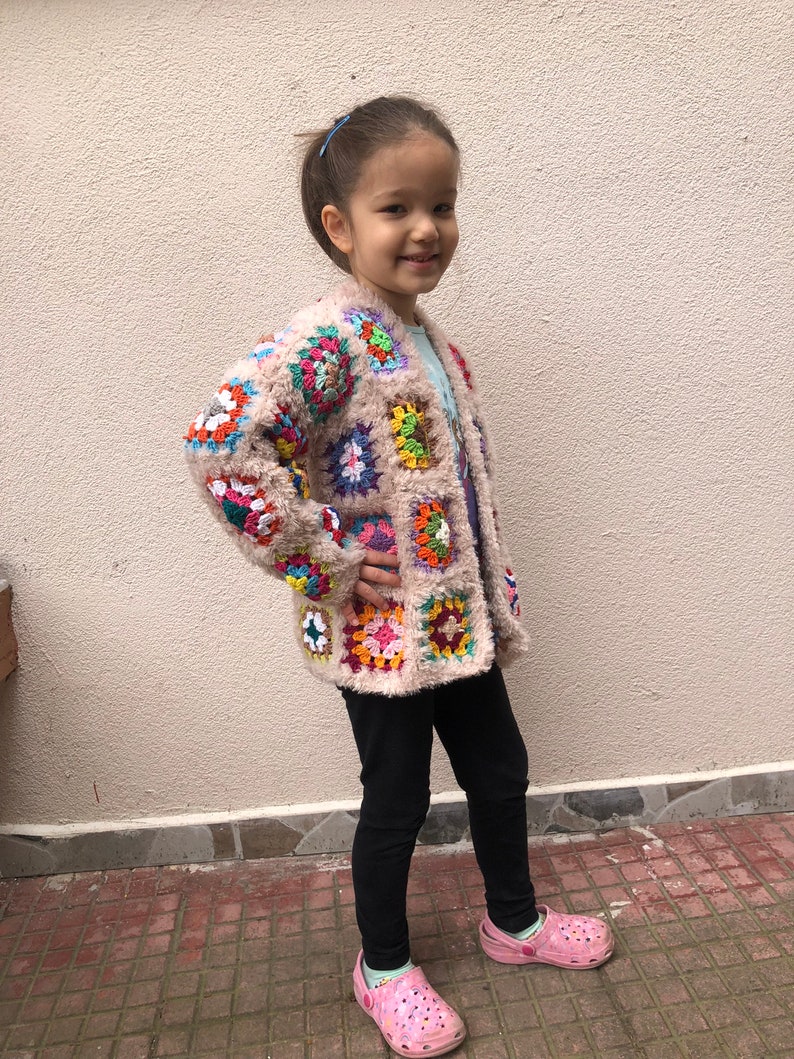 May include: A young person wearing a colorful crocheted cardigan with a fuzzy texture. The cardigan features a variety of colors, including pink, blue, green, yellow, and orange. The person is wearing black leggings and pink and green shoes.