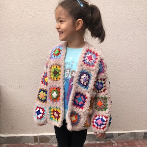 May include: A young girl wearing a colorful crocheted cardigan with a fuzzy texture. The cardigan features a variety of colors, including red, orange, yellow, green, blue, purple, and pink. The girl is wearing a blue shirt with a Frozen character design and black leggings.