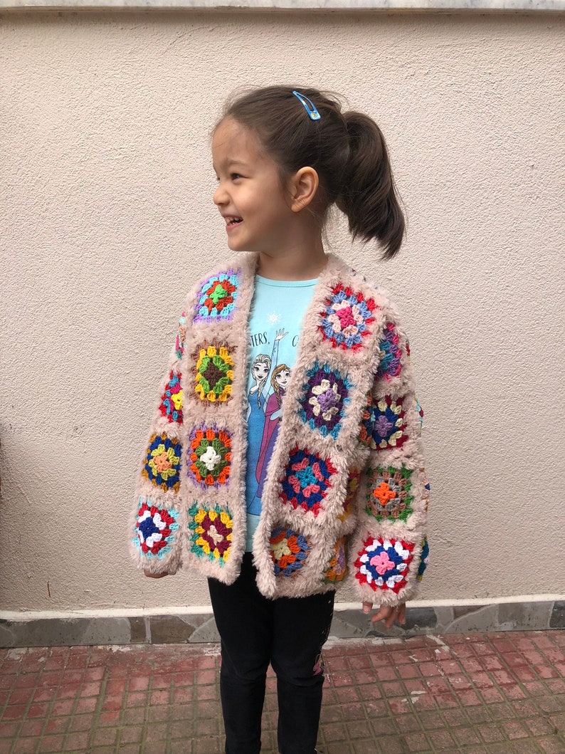 May include: A young girl wearing a colorful crocheted cardigan with a fuzzy texture. The cardigan features a variety of colors, including red, orange, yellow, green, blue, purple, and pink. The girl is wearing a blue shirt with a Frozen character design and black leggings.