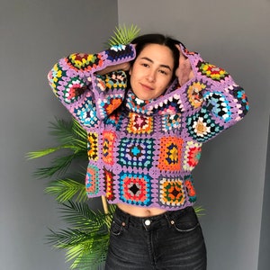 May include: A colorful crocheted sweater with a high neck and a geometric pattern. The sweater is made of a variety of colors, including purple, pink, blue, green, yellow, and orange.