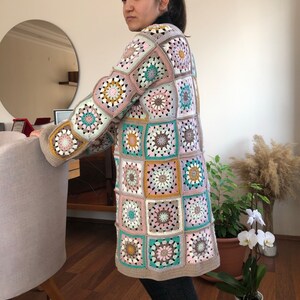 Könnte beinhalten: Ein langer, gehäkelter Cardigan mit Patchwork-Muster aus Quadraten in verschiedenen Farben, darunter Rosa, Grün, Braun und Beige.