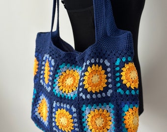May include: A blue crocheted tote bag with a colorful geometric pattern. The bag has two long straps and is made of a soft, textured yarn.