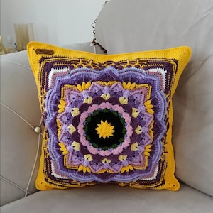 May include: A square, handmade crochet cushion with a vibrant mandala design. The cushion features a yellow border and a central pattern in shades of purple, lavender, and yellow. The centre of the mandala is black with a yellow starburst.