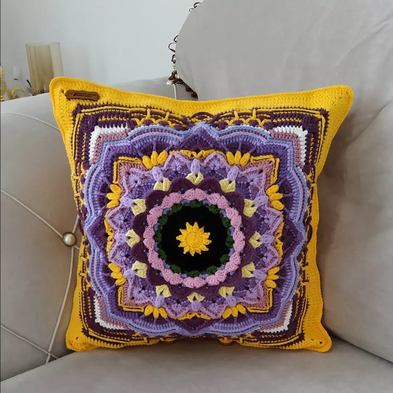 May include: A square, handmade crochet cushion with a vibrant mandala design. The cushion features a yellow border and a central pattern in shades of purple, lavender, and yellow. The centre of the mandala is black with a yellow starburst.