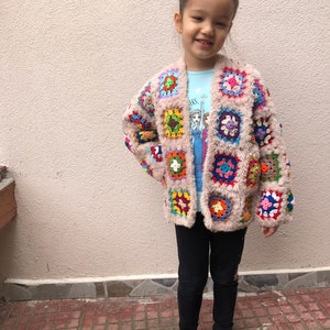 May include: A young girl wearing a pink crocheted cardigan with colorful squares. The cardigan has a fuzzy texture and is open in the front. The girl is wearing black leggings and pink shoes.