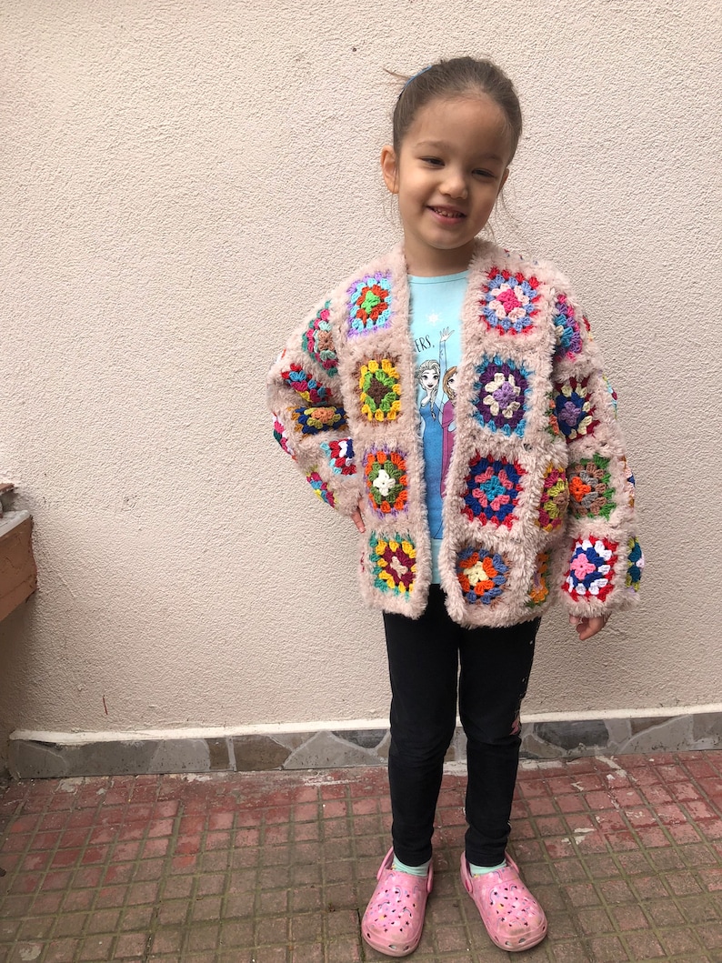 May include: A young girl wearing a pink crocheted cardigan with colorful squares. The cardigan has a fuzzy texture and is open in the front. The girl is wearing black leggings and pink shoes.