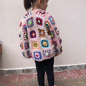 May include: A child wearing a pink crocheted cardigan with colorful square patches. The cardigan has a fuzzy texture. The child is wearing black leggings and pink shoes.
