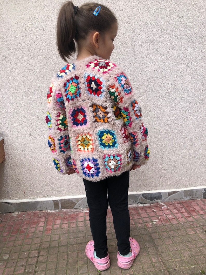 May include: A child wearing a pink crocheted cardigan with colorful square patches. The cardigan has a fuzzy texture. The child is wearing black leggings and pink shoes.