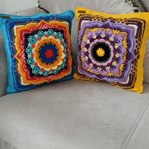 May include: Two square crochet cushions with detailed mandala patterns. One has a blue border with an orange, red, and yellow central design. The other has a yellow border with a purple and green mandala design. Both cushions are on a grey sofa.