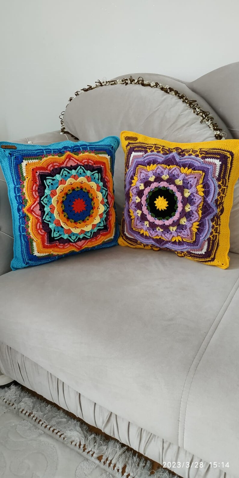 May include: Two square crochet cushions with detailed mandala patterns. One has a blue border with an orange, red, and yellow central design. The other has a yellow border with a purple and green mandala design. Both cushions are on a grey sofa.