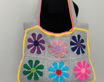 May include: A gray crocheted tote bag with a yellow and pink border. The bag features six colorful flower designs in shades of purple, pink, blue, green, and red.