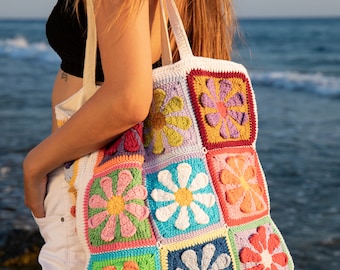 May include: A colorful crocheted tote bag with a daisy pattern. The bag has a white canvas strap and fringe trim.