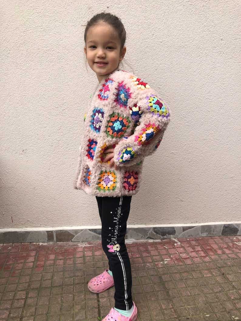 May include: A young person wearing a pink crocheted cardigan with colorful squares and black leggings with white stripes and the text "Minnie Mouse".