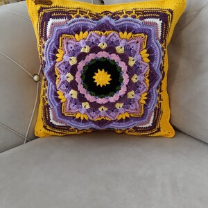 May include: A square cushion with a crocheted mandala design. The cushion has a yellow border and a central pattern in shades of purple, yellow, and black. The cushion is on a grey sofa.