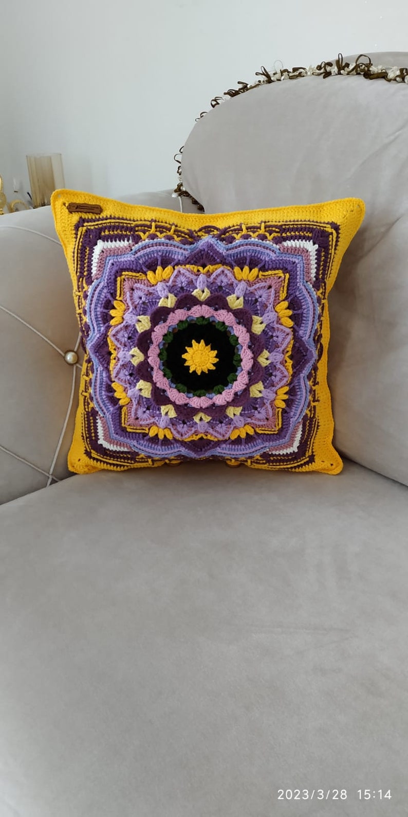 May include: A square cushion with a crocheted mandala design. The cushion has a yellow border and a central pattern in shades of purple, yellow, and black. The cushion is on a grey sofa.