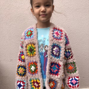May include: A child wearing a light pink cardigan with colorful crocheted squares. The cardigan has a fuzzy texture and is open in the front. The child is wearing a blue shirt with a snowflake design and black pants.