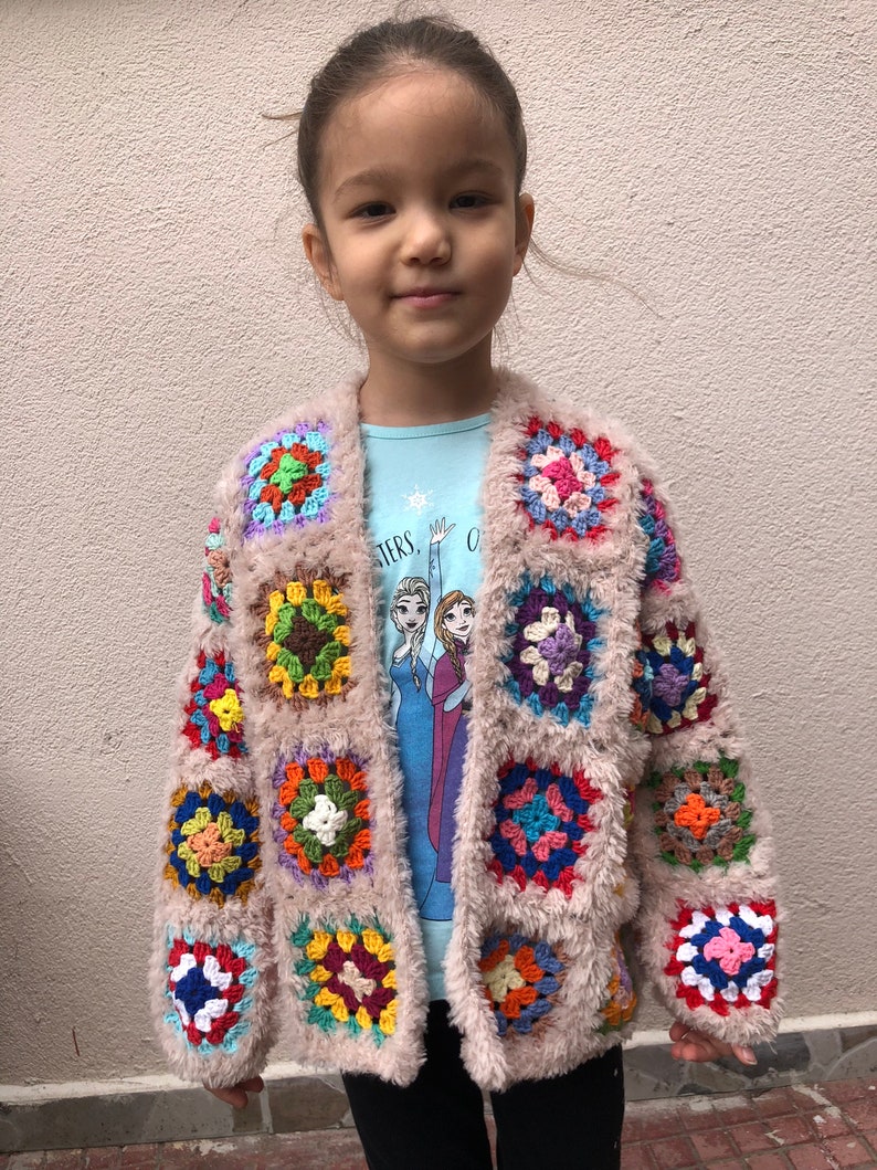 May include: A child wearing a light pink cardigan with colorful crocheted squares. The cardigan has a fuzzy texture and is open in the front. The child is wearing a blue shirt with a snowflake design and black pants.