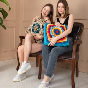 May include: Two square crochet cushions. One features a beige and dark blue geometric design. The other has a vibrant mandala pattern in turquoise, red, orange, yellow, and blue. The cushions are decorative and handmade.