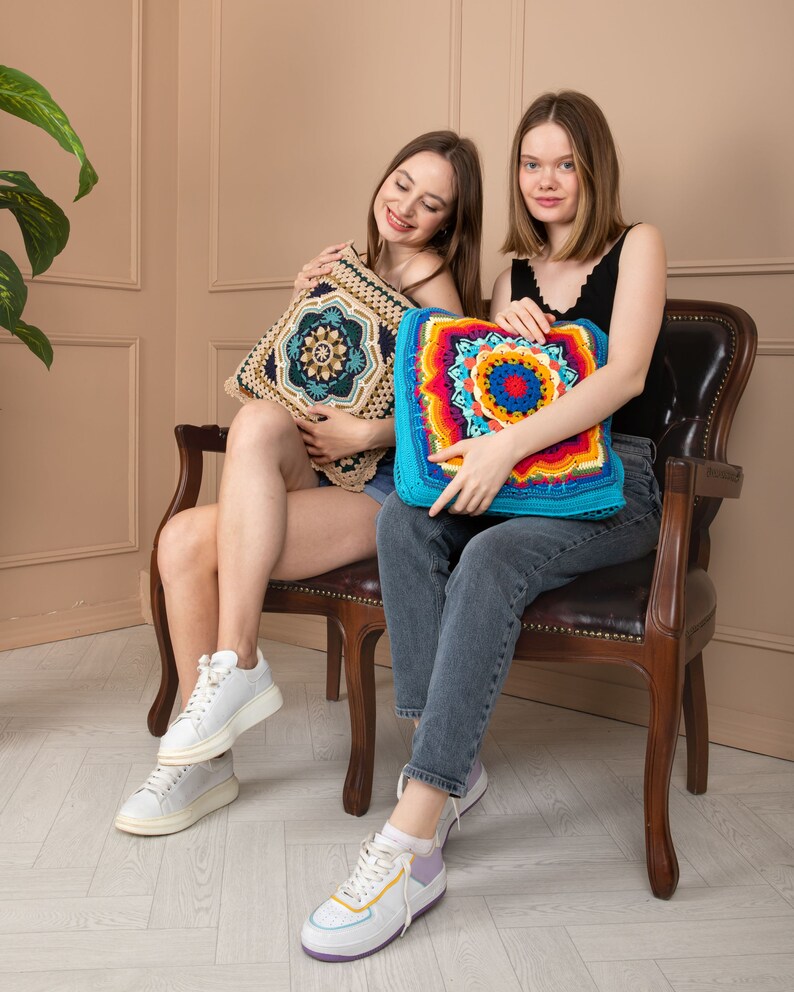 May include: Two square crochet cushions. One features a beige and dark blue geometric design. The other has a vibrant mandala pattern in turquoise, red, orange, yellow, and blue. The cushions are decorative and handmade.