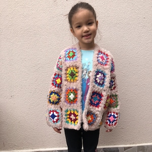 May include: A child wearing a light brown crocheted cardigan with colorful square patches. The cardigan has a fuzzy texture and is worn over a blue shirt with a cartoon image. The child is wearing black pants.