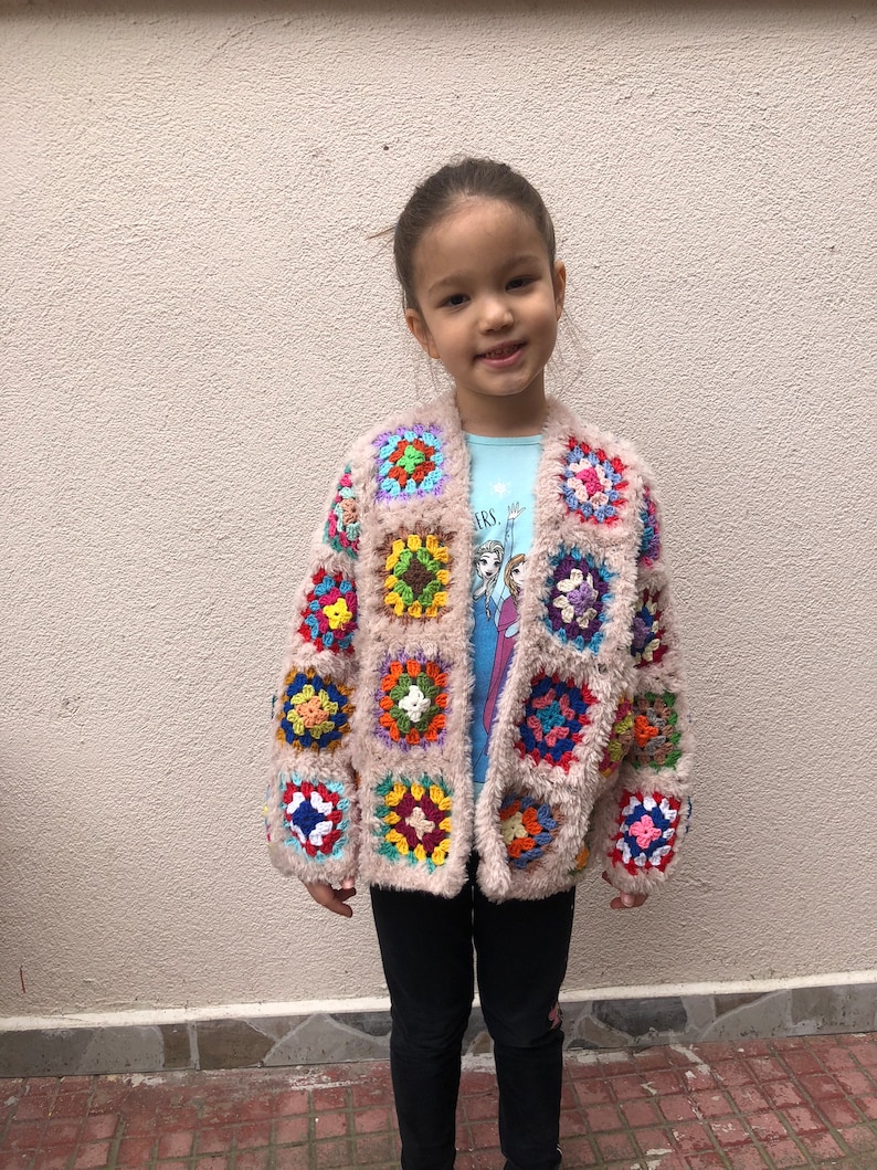 May include: A child wearing a light brown crocheted cardigan with colorful square patches. The cardigan has a fuzzy texture and is worn over a blue shirt with a cartoon image. The child is wearing black pants.