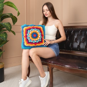 May include: A square, crocheted cushion with a vibrant mandala design. The cushion features a turquoise border, with concentric circles of orange, yellow, red, and blue. The cushion is held by a person wearing a white vest and denim shorts.