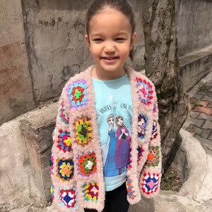 May include: A young girl wearing a pink crocheted cardigan with colorful squares. The cardigan has a fuzzy texture. The girl is wearing a light blue t-shirt with a Frozen image and the text "Two Sisters One Heart". She is also wearing black pants.