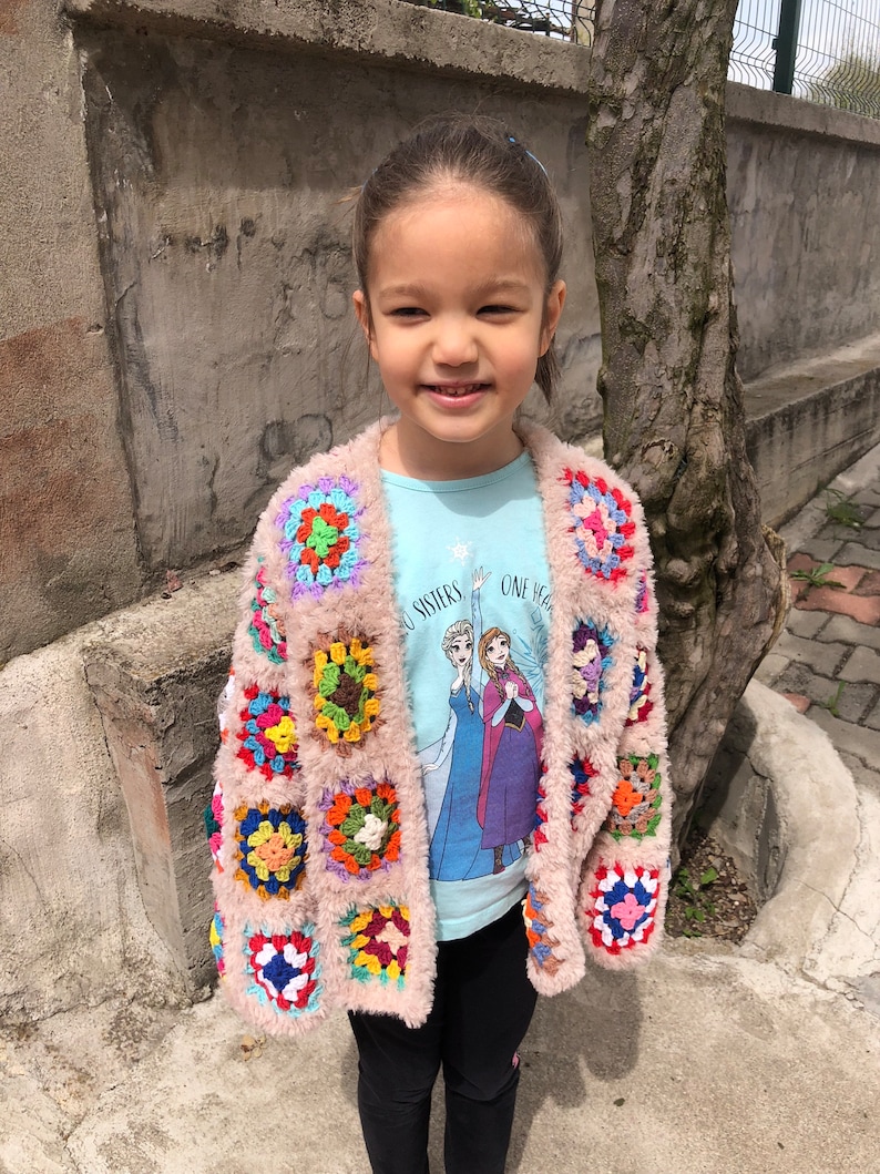May include: A young girl wearing a pink crocheted cardigan with colorful squares. The cardigan has a fuzzy texture. The girl is wearing a light blue t-shirt with a Frozen image and the text "Two Sisters One Heart". She is also wearing black pants.