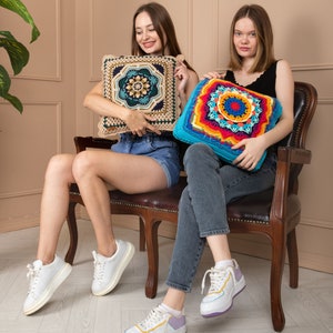 May include: Two square crochet cushions. One is beige with a dark teal and navy blue geometric design. The other is turquoise with a vibrant rainbow mandala pattern. Both cushions are held by people sitting on a brown leather chair.