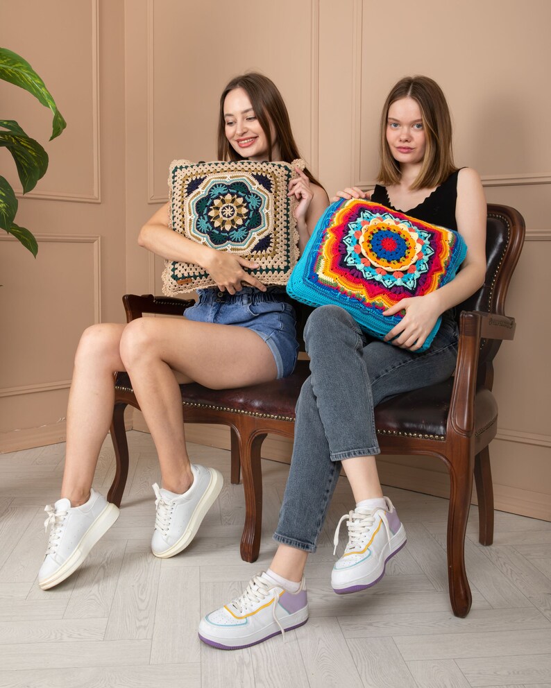 May include: Two square crochet cushions. One is beige with a dark teal and navy blue geometric design. The other is turquoise with a vibrant rainbow mandala pattern. Both cushions are held by people sitting on a brown leather chair.
