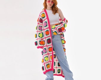 May include: A long white crochet cardigan with pink trim and colorful flower embellishments. The cardigan is open in the front and has a relaxed fit.
