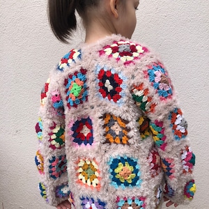 May include: A pink fuzzy cardigan with a colorful crocheted design. The cardigan is made up of squares of different colors and patterns.