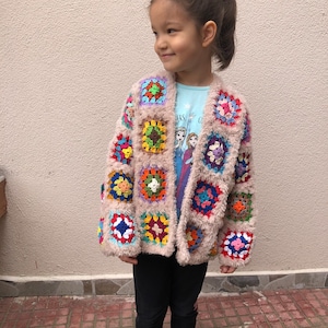 May include: A young girl wearing a beige crocheted cardigan with colorful square patches. The cardigan has a fuzzy texture and is open in the front. The girl is wearing black pants and pink shoes.