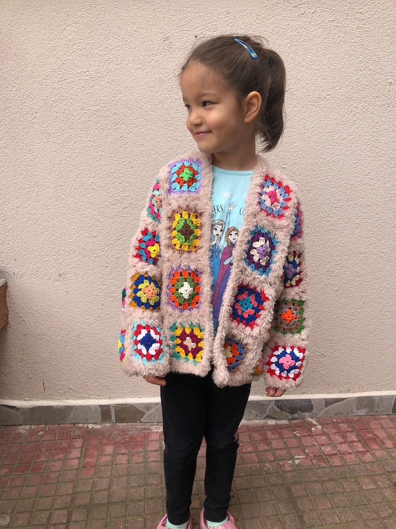 May include: A young girl wearing a beige crocheted cardigan with colorful square patches. The cardigan has a fuzzy texture and is open in the front. The girl is wearing black pants and pink shoes.