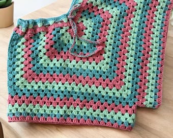 May include: A crocheted garment with a geometric pattern in shades of pink, green, and blue. The garment has a drawstring at the top and is made with a granny square stitch.