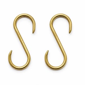Peut inclure: Deux crochets en forme de S identiques fabriqués en métal laiton poli et lisse. Chaque crochet présente un design incurvé continu avec des extrémités arrondies, formant une forme symétrique permettant de suspendre des objets.