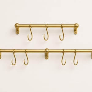 Può includere: Due barre orizzontali da parete realizzate in ottone spazzolato. La barra più corta presenta tre ganci a forma di S, mentre quella più lunga ne include quattro. Entrambe le barre hanno terminali arrotondati e piastre di fissaggio con viti.