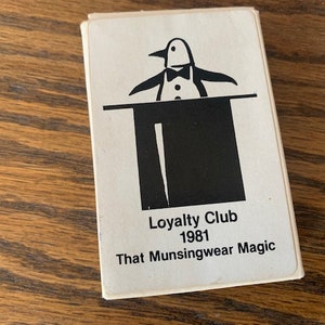 May include: A white card with a black and white illustration of a penguin wearing a bow tie standing on a black top hat. The text on the card reads "Loyalty Club 1981 That Munsingwear Magic".