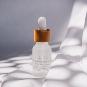 Może przedstawiać: Przezroczysta butelka ze szkła z bambusową nakrętką i kroplomierzem, zawierająca serum z etykietą "Gentle Niacinamide Serum, protecting and soothing". Butelka stoi na białej powierzchni z tkaniny.
